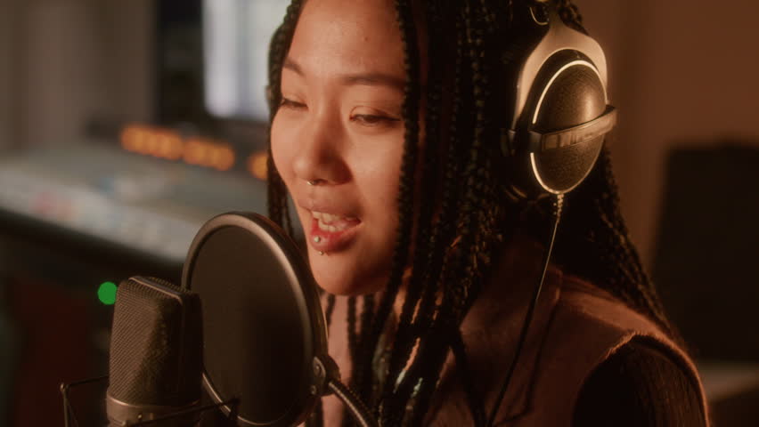 Young Asian female singer with long braids and headphones singing into professional condenser microphone during vocal recording session in music studio. Close up shot