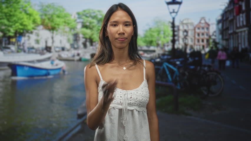 Thai woman in white dress holding cigarette to lips, fingers near mouth, facing camera on a street beside a canal and parked bicycles; quiet contemplation.