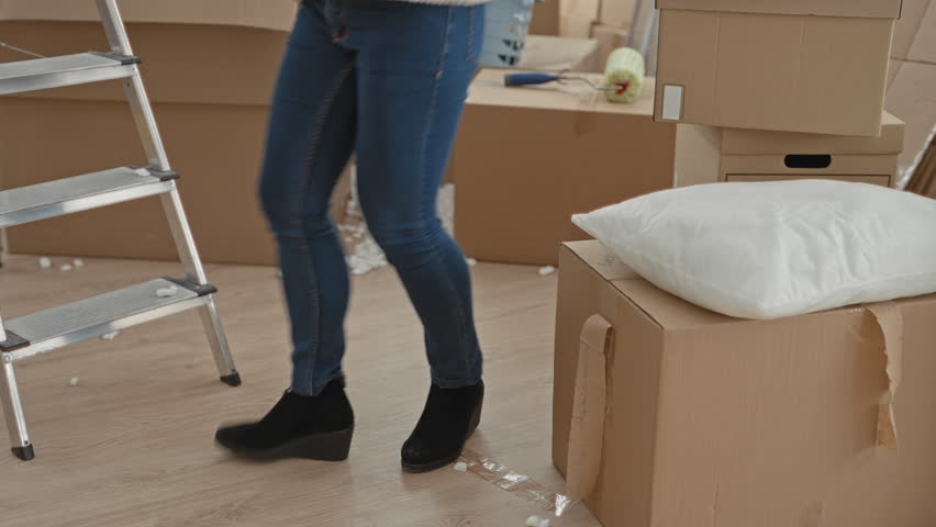 Woman in jeans and black wedge boots walking between packed cardboard boxes, ladder and pillow in an apartment during a move; moving day focused.