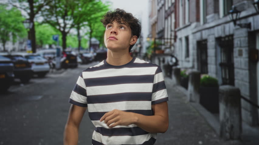 Teenage boy pointing finger upward, arms crossed, standing on a city street by parked cars and stone building; quiet curiosity.
