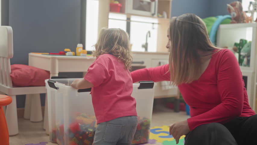 Woman kneeling by a clear plastic toybox helps small girl reach colorful blocks and holds a red toy in building playroom; parental care calm.