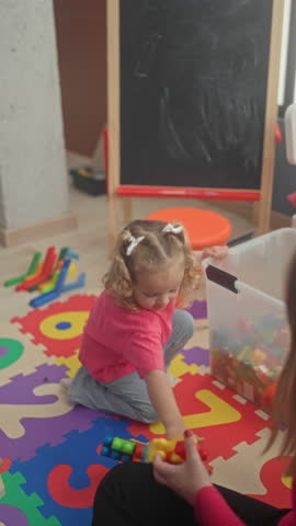 Little girl sitting and playing with colorful toy blocks on a foam puzzle mat next to a chalkboard easel in a preschool building; early learning joy.
