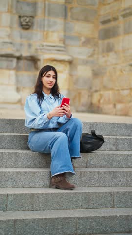 Stylish young woman sitting on the historic stone steps of a church, smiling and taking selfies with her red smartphone