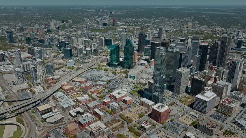 A sweeping aerial perspective of the Dallas, Texas metropolitan area. The footage captures the dense concentration of modern skyscrapers in the central business district, including the distinctive green glass of Fountain Place and the towering Bank of America Plaza. The shot highlights the complex urban grid, including major highway arteries and city transit lines. Ideal for commercial real estate marketing, corporate videos, and regional documentaries focusing on Texas urban development and metropolitan growth.
