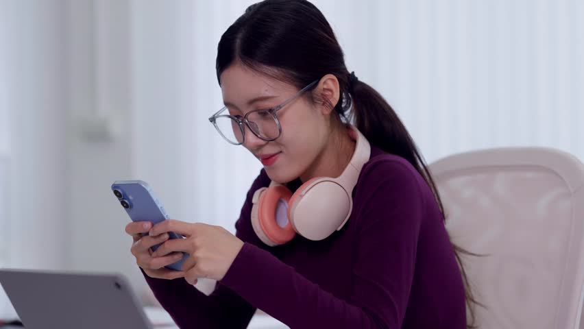 young Asian women using smartphone texting message cheerful smile with headphones laptop computer hybrid remote work