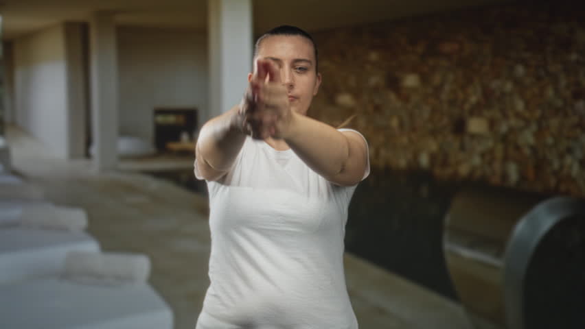 Woman in white t shirt making a finger gun gesture with hands pointed forward and one eye closed in a building corridor with lounge beds; determination focus.