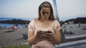Woman frowning and holding a smartphone with both hands on a beach, staring at the screen while tapping with thumbs; digital frustration. - Powered by Shutterstock - Get 15% off with code: PIKWIZARD15