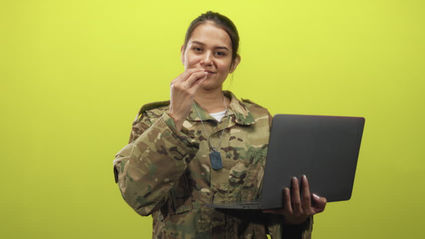 Woman soldier in camouflage uniform holds a laptop in one hand and makes a finger to lips gesture while looking aside in studio; pride duty.