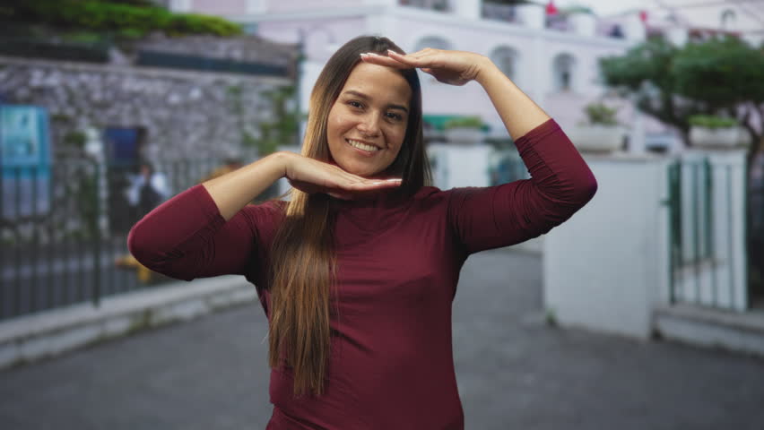Woman young hispanic brunette smiling framing her face with hands near temples on street by a stone wall and iron fence; playful joy.