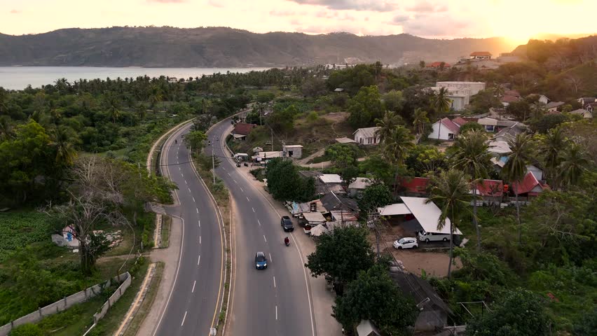 Picturesque aerial view of a winding coastal road with cars and motorcycles driving through a lush tropical landscape with the ocean and mountains in the background during a beautiful golden sunset