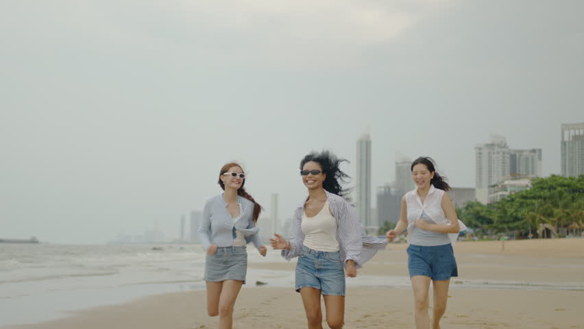 Three diverse young women running and jumping joyfully on a sandy beach with a city skyline background. Happy multicultural female friends enjoying a sunny summer vacation and freedom by the ocean