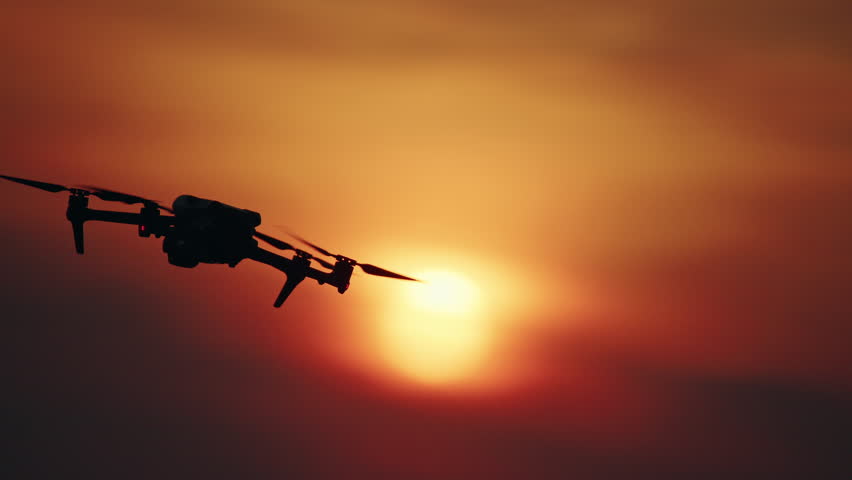 Drone silhouette flying across glowing sunset sun with orange sky.