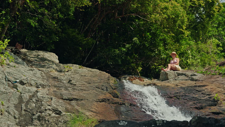 Young woman sitting on rocky ledge beside small waterfall in tropical jungle, enjoying quiet moment of travel and nature in green mountain landscape. High quality 4k footage