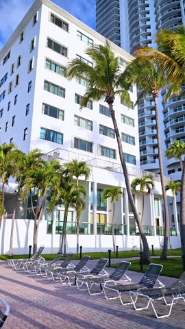 Lounge chairs by white hotel building and palm trees. Outdoor view of a row of beach chairs on a patio next to a white modern building and palm trees in Miami Beach.