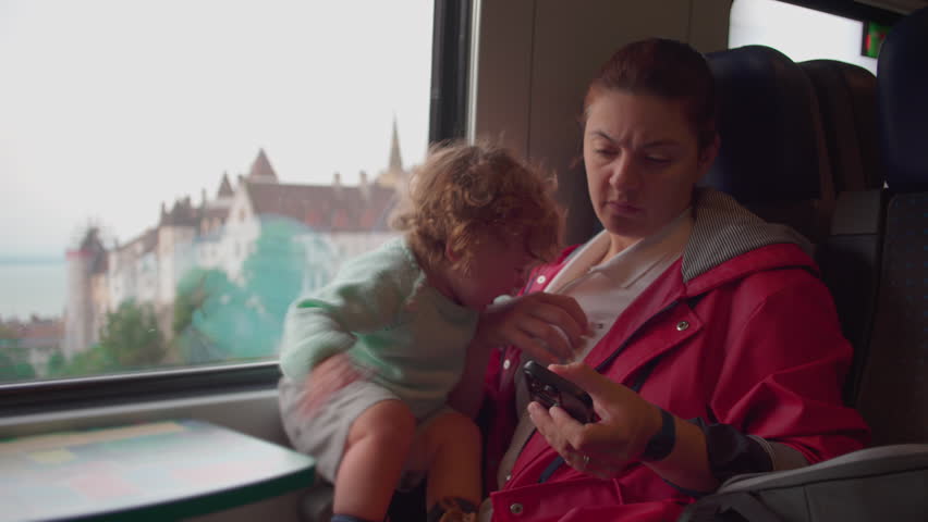 Mother looking at smartphone while toddler reaches for attention on moving train with blurred view of European town and church spires through window