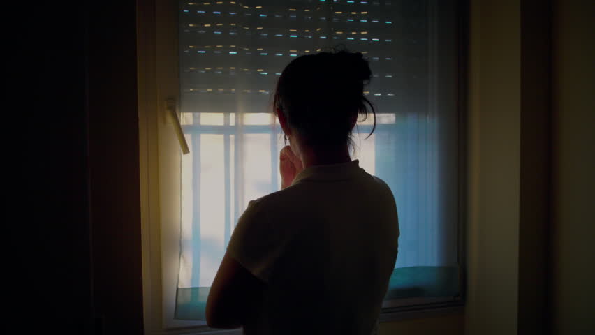 Rear view of woman standing in bedroom facing window, sheer curtain filtering light as she looks outside with a worried stance