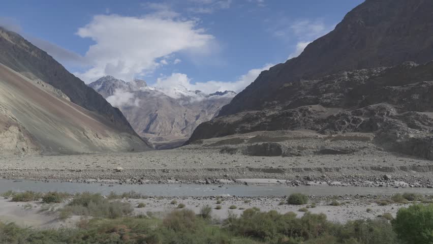Cinematic aerial drone movement gliding right to left over a flowing river in Ladakh, with layered Himalayan mountains and clouds quietly resting over the high peaks creating a dramatic landscape.