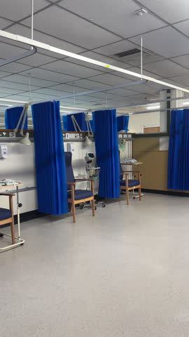 Vertical. Panning shot of empty cubicles in hospital ward. Blue patient privacy curtains hanging open in an open space.