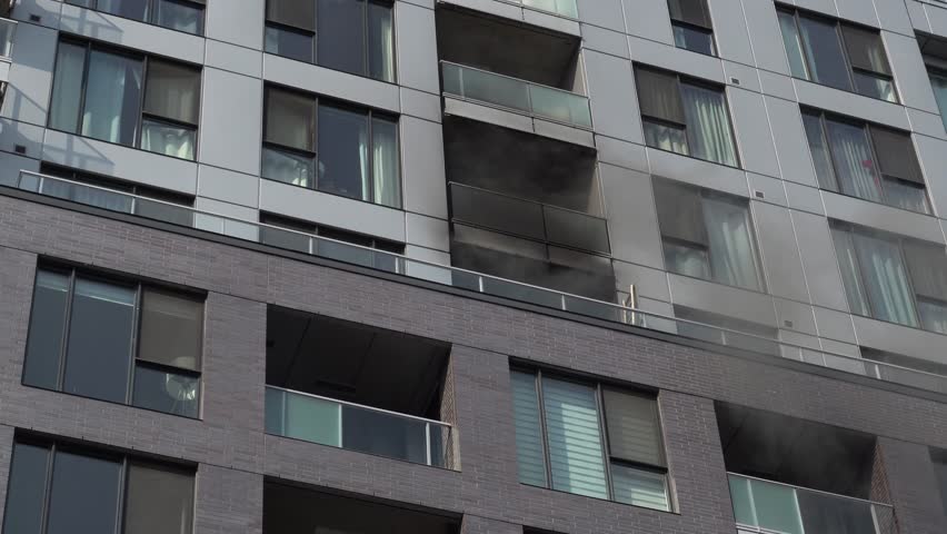 Low-angle shot of a modern residential skyscraper showing charred balconies and smoke damage on the gray facade following a fire. High-resolution architectural assessment.