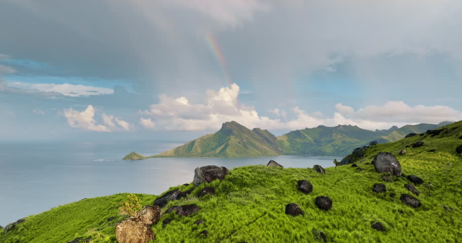 Lush green hills and volcanic mountains in the Fiji islands rising from calm turquoise ocean waters under a cloudy sky with a vibrant rainbow arching over the distant peaks