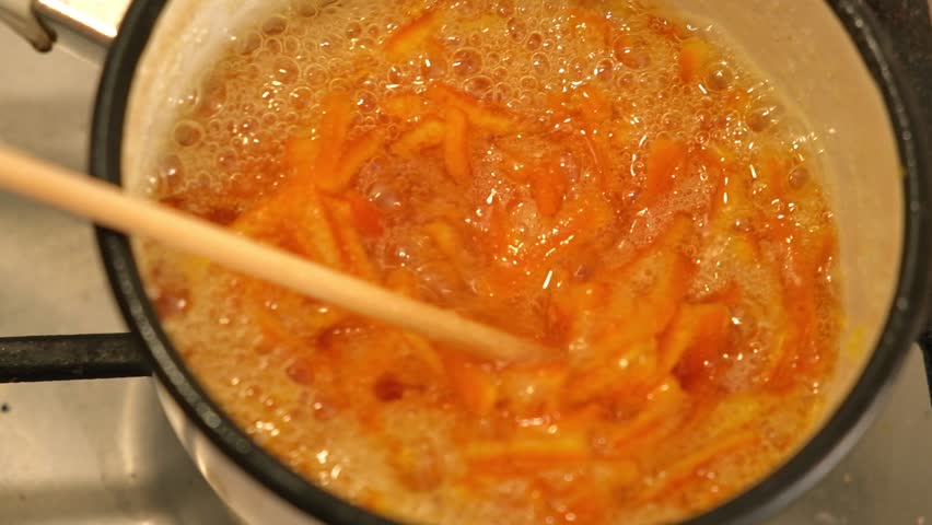 Candied orange peels simmering in pot, chopstick stirring glossy citrus strips in bubbling sugar syrup. warm kitchen light. Smallbatch handmade confection process, sticky caramelizing. Citrus sweets
