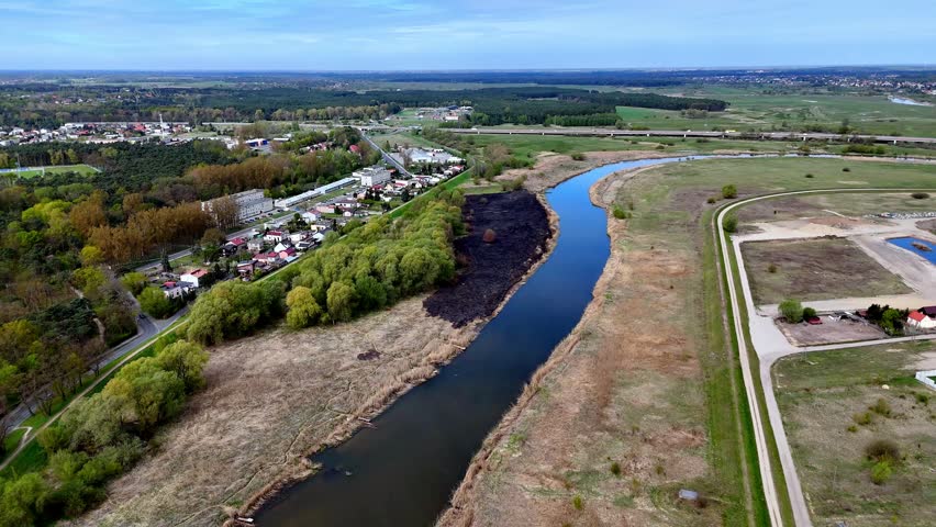 Aerial drone view of a river bend featuring a large black scorched earth area after a grass fire, adjacent to lush green trees, suburban housing, a highway bridge, and meadows.