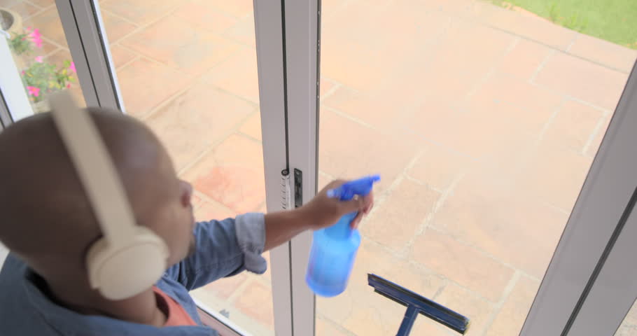 African man approaching sliding door, holding blue spray, cleaning glass with squeegee, copy space. Window, patio, plants, terrace, denim, home, wash