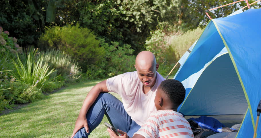 African father and son sitting on grass by blue tent adult leaning explaining phone youth reaching. Camping, outdoor, lawn, smartphone, learning, bonding, candid
