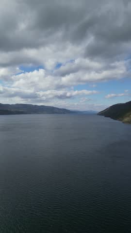A static aerial view of a calm lake surrounded by soft rolling hills under a cloudy sky