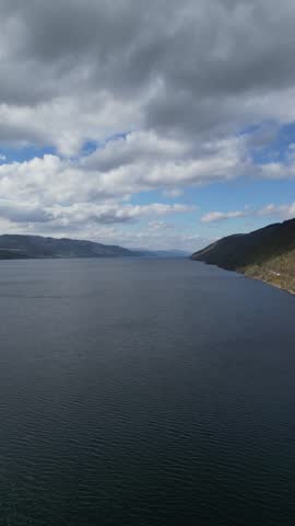 A static aerial view of a tranquil lake surrounded by rolling hills under a soft cloudy sky