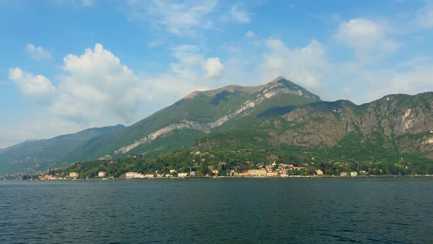 Wide view of Lake Como with colorful coastal town at the foot of massive green mountain under blue sky with clouds in Italy.