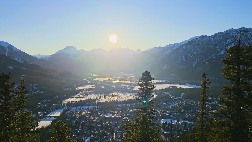 Sunset aerial-style view of Banff valley in the Canadian Rockies, featuring snow covered fields, mountain ridges, forest, town buildings, glowing alpine scenery, and winter tourism.
