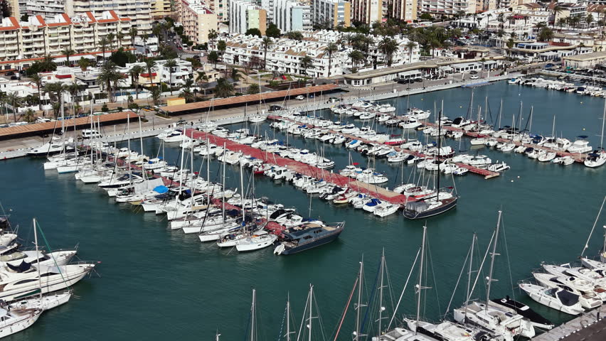 Many boats are parked at a marina with buildings and trees in the background. The scene shows the hustle of a busy coastal town during daytime.
