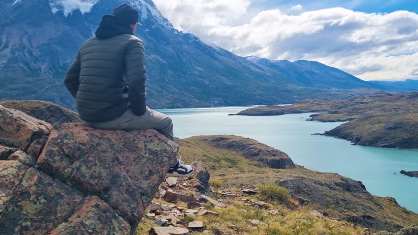 A man sits on a rock overlooking the wild Patagonian landscape of Torres del Paine National Park, Chile, with Lake Pehoé below on a partly cloudy day, taking in the vast mountain scenery