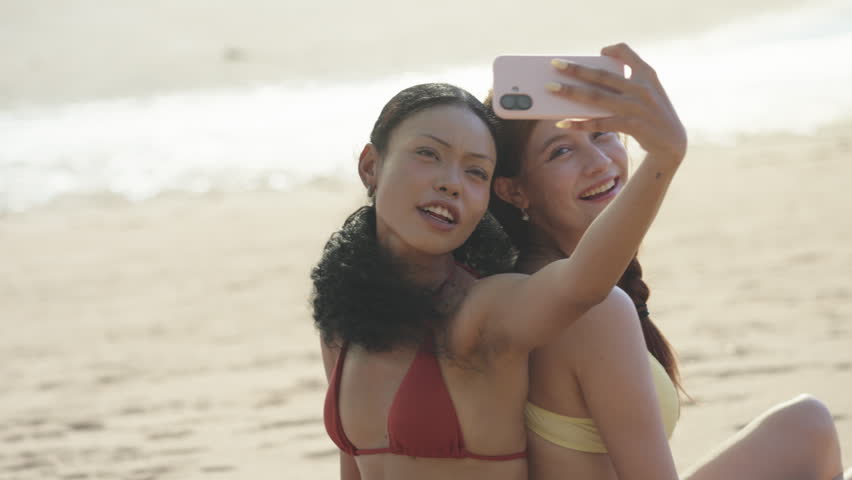Two happy multiethnic young women in bikinis smiling while taking a selfie with a smartphone on a sunny tropical beach during their summer vacation travel at sunset time outdoors.