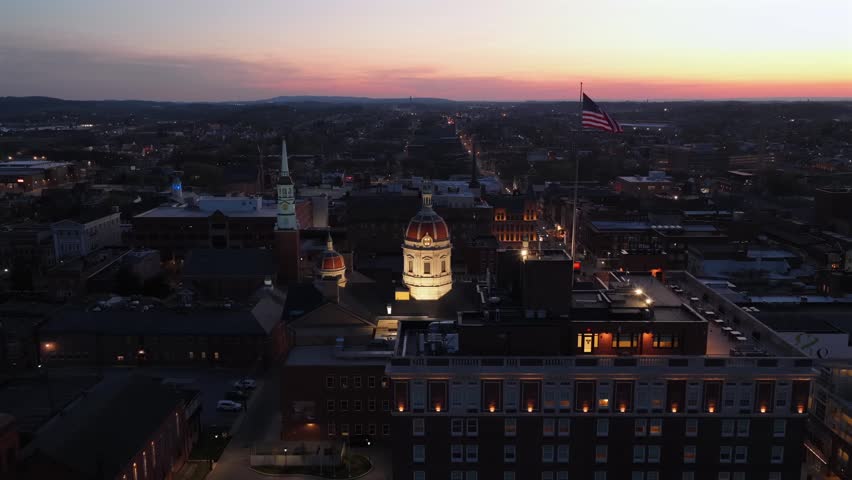 Aerial establishing shot of downtown York, Pennsylvania at sunset with illuminated county courthouse dome, waving American flag, historic skyline and warm evening twilight over city in USA. Wide shot.