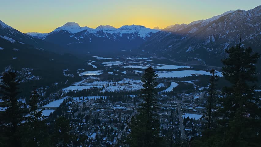 Golden sunset view across Banff, Canada, featuring snow covered mountains, pine forest, frozen valley, town buildings, glowing sky, scenic Rocky Mountain destination, and winter travel.