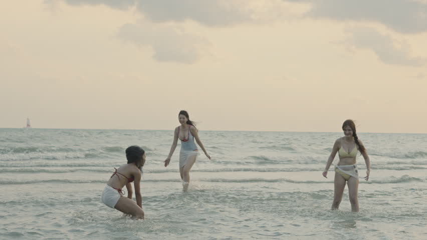 Three happy multiethnic young women in swimwear having fun and splashing water while relaxing on a beautiful tropical beach during their summer vacation travel at sunset time.