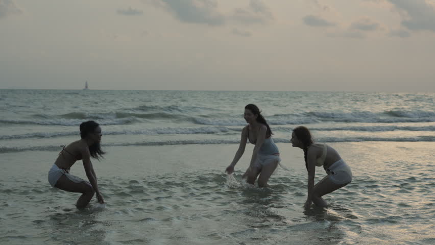 Three happy multiethnic young women in swimwear having fun and splashing sea water together while enjoying a beautiful sunset on a tropical beach during their summer vacation travel.