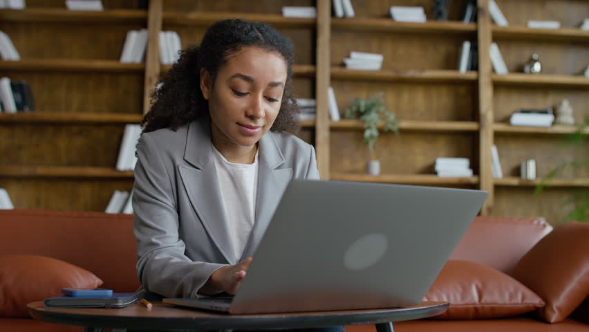 A young professional woman works on a laptop in a cozy office lounge with bookshelves and warm decor