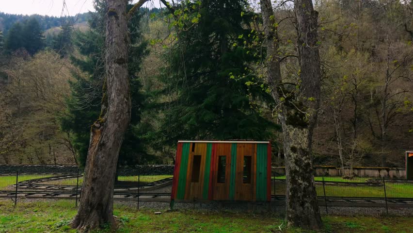 Small wooden hut painted in vibrant green and red stands beside narrow-gauge railway tracks within a forested park. Tall trees frame the scene under a soft cloudy sky, highlighting nature. childhood.