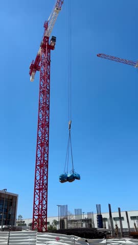 Red tower crane lifting heavy equipment over concrete structure framework against clear blue sky at construction site
