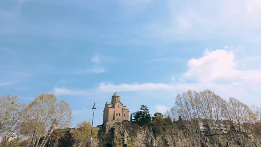 Iconic Metekhi Church perched atop a steep rocky cliff, under a vast blue sky with light clouds. Trees with early spring foliage surround the historic landmark in Tbilisi.