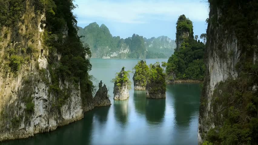 Towering limestone karsts covered in dense vegetation emerge from a calm, reflective turquoise lake. Distant, hazy green mountains form the backdrop under a clear blue sky.