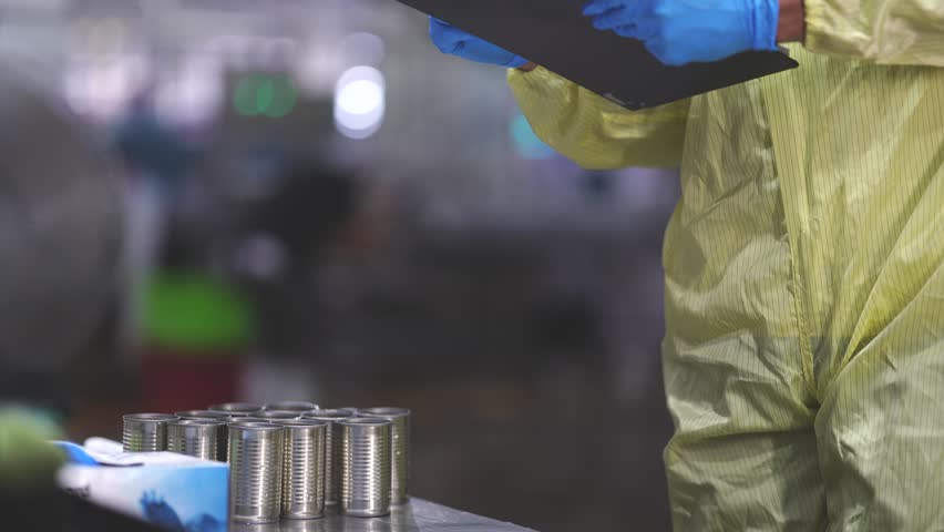 A professional female quality controller in a sanitized lab coat and safety glasses inspecting an automated production line in a modern manufacturing facility.