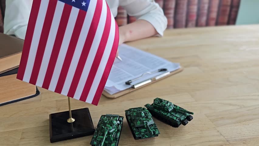 Toy tanks are positioned by a US flag on a wooden table. A person is writing on papers and using a clipboard. The setting shows a mix of play and work.