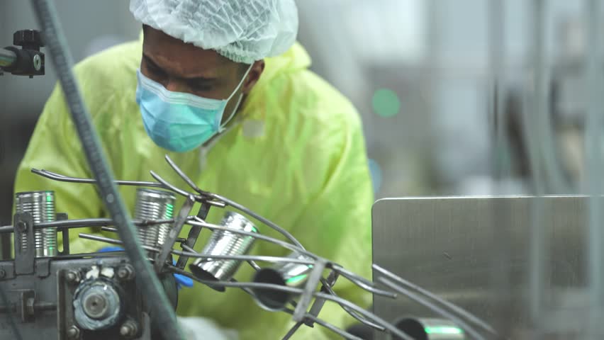 A focused industrial auditor in a yellow protective suit and mask recording quality control data while overseeing production in a modern manufacturing plant.