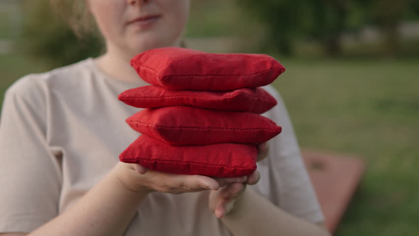 Impersonal woman holding stack of red cornhole bags in hands for cornhole game. Female player smiling while looking at set of sports equipment for friendly popular american outdoor game.