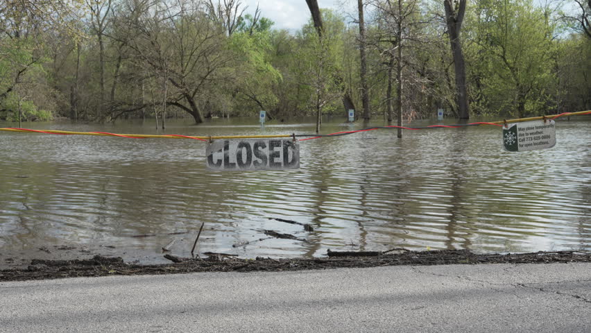 Flooded park area and submerged parking lot after heavy rainfall. Water levels rose across the area, forcing closure signs near the flooded roadway and trees.