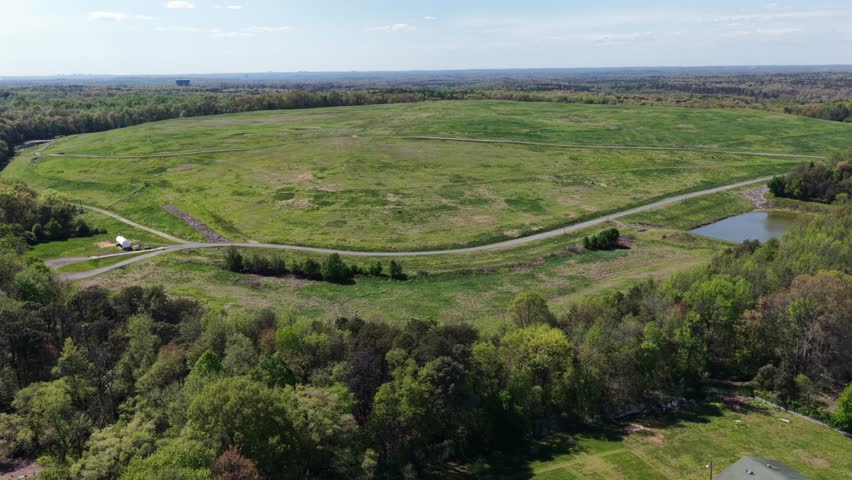 Expansive rural farmland landscape with winding dirt road, green fields and small pond surrounded by trees showing peaceful countryside scenery in United States during spring season. Aerial wide view.